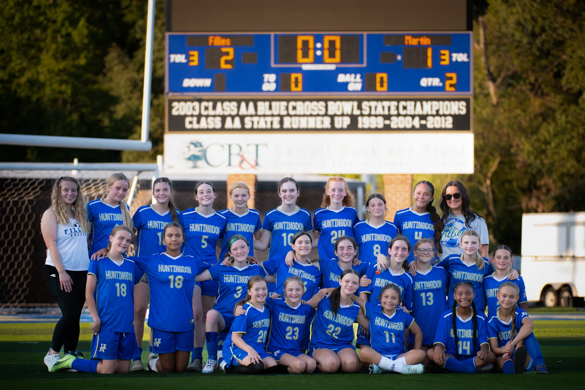 Full girls soccer squad and coaches lined up on the field beneath the scoreboard after a win