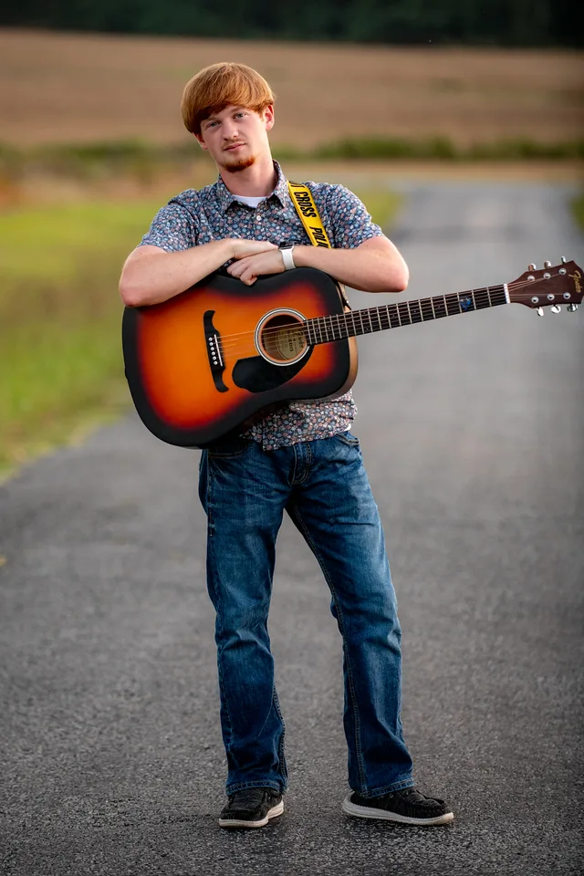Senior portrait of a young man standing with his guitar on a country road at sunset